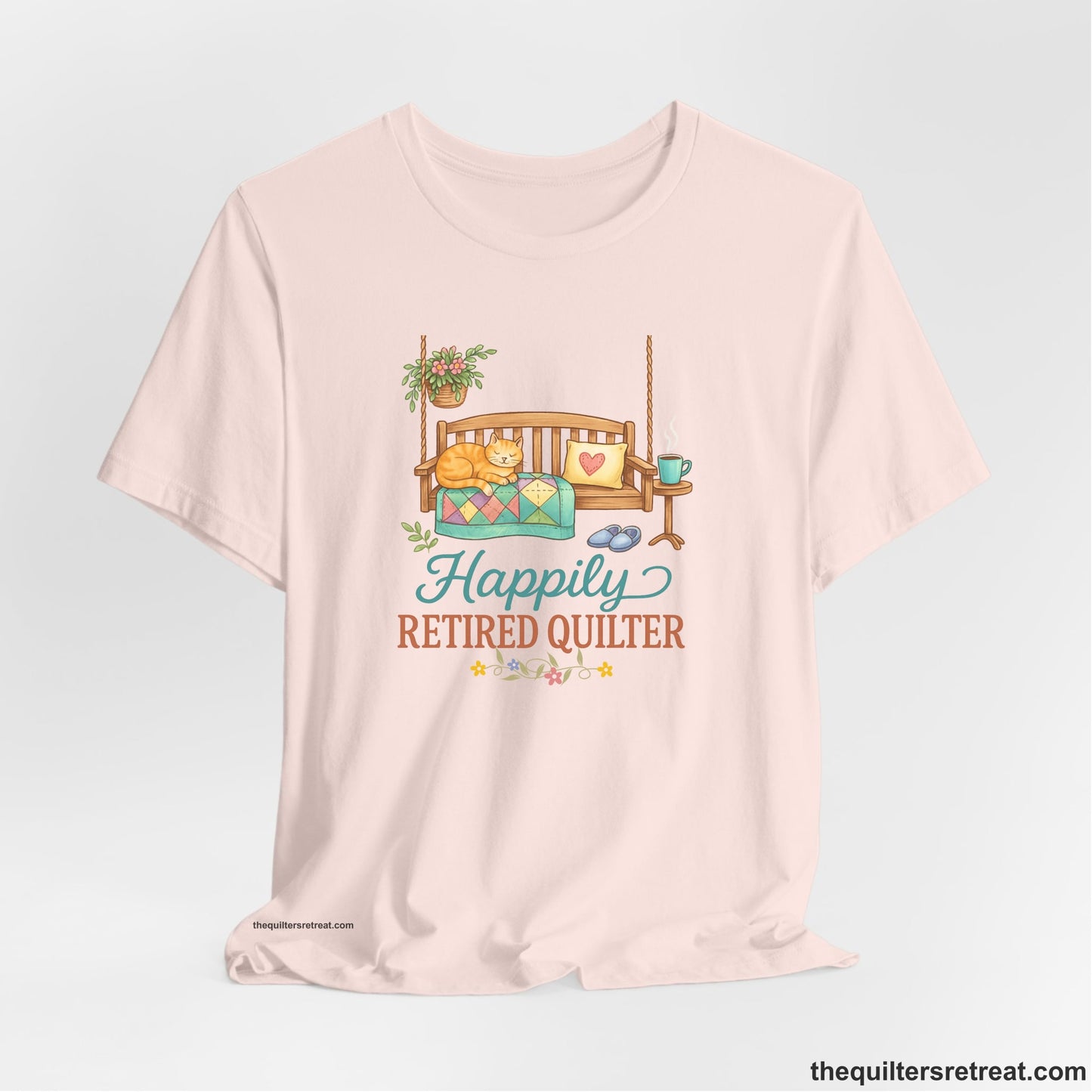 A Soft Pink Short Sleeve T-Shirt with a hanging bench with cozy cat and quilt, surrounded by plants, "Happily Retired Quilter" text